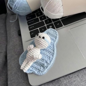Op de afbeelding: Een handgemaakte witte gehaakte babypop met zwarte ogen en een blauwe streep op het hoofd, liggend op een lichtblauwe gehaakte deken. De pop ligt op een laptoptoetsenbord, met twee bollen wol op de achtergrond.