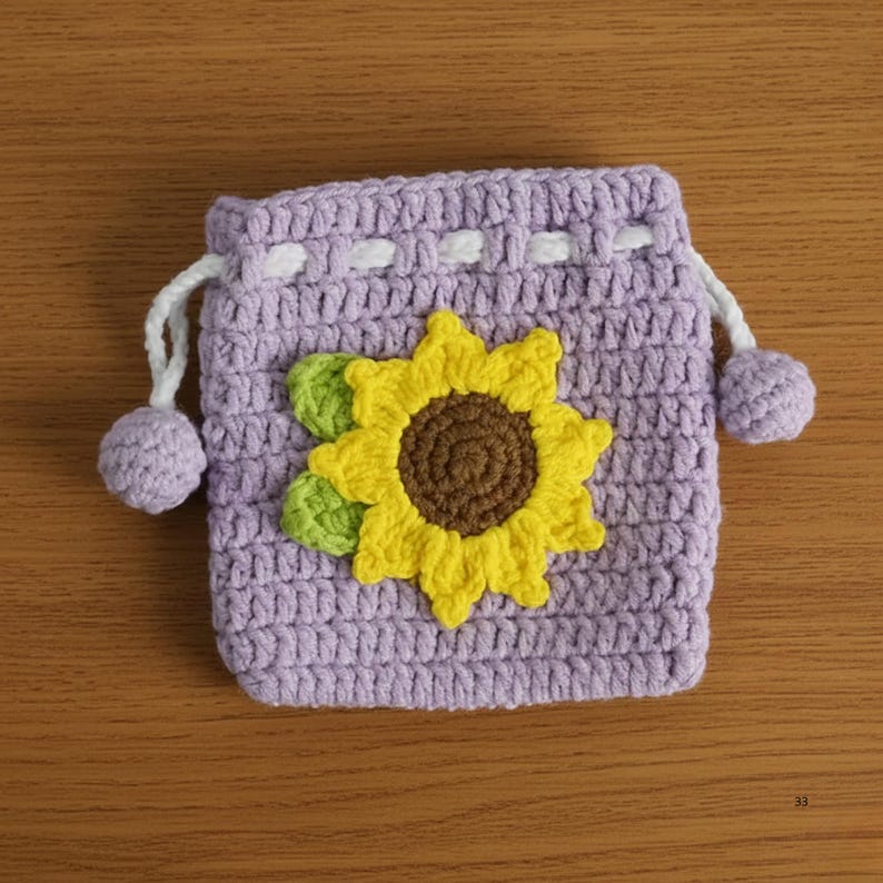 K&ouml;nnte beinhalten: Ein geh&auml;kelter, lavendelfarbener Kordelzugbeutel mit einer Applikation einer gelben Sonnenblume. Die Sonnenblume hat eine braune Mitte und gr&uuml;ne Bl&auml;tter. Der Beutel hat wei&szlig;e Kordeln und lavendelfarbene Pompons.