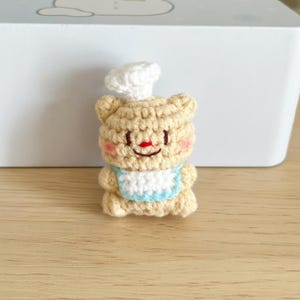 May include: A crocheted bear figurine wearing a chef's hat and a blue-trimmed bib. The bear is tan with a red nose and pink cheeks. The figurine is handmade and sits on a wooden surface.