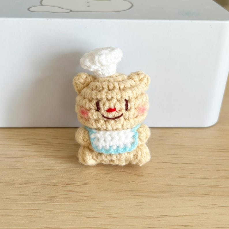 May include: A crocheted bear figurine wearing a chef's hat and a blue-trimmed bib. The bear is tan with a red nose and pink cheeks. The figurine is handmade and sits on a wooden surface.