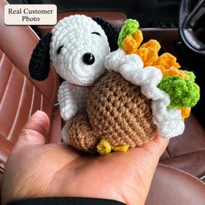 May include: A white and black crocheted dog figure with closed eyes and a small red collar, holding a brown crocheted acorn-shaped object topped with white frosting, orange textured elements, and green crocheted leaves.