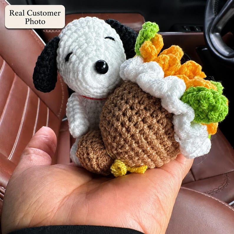 May include: A white and black crocheted dog figure with closed eyes and a small red collar, holding a brown crocheted acorn-shaped object topped with white frosting, orange textured elements, and green crocheted leaves.