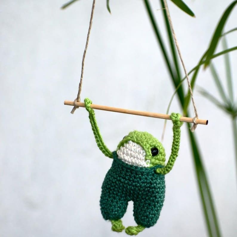 May include: A handmade crochet frog toy in shades of green and white, hanging from a wooden bar. The frog is wearing green overalls and has black eyes. The toy is suspended by twine, creating a whimsical, decorative piece.