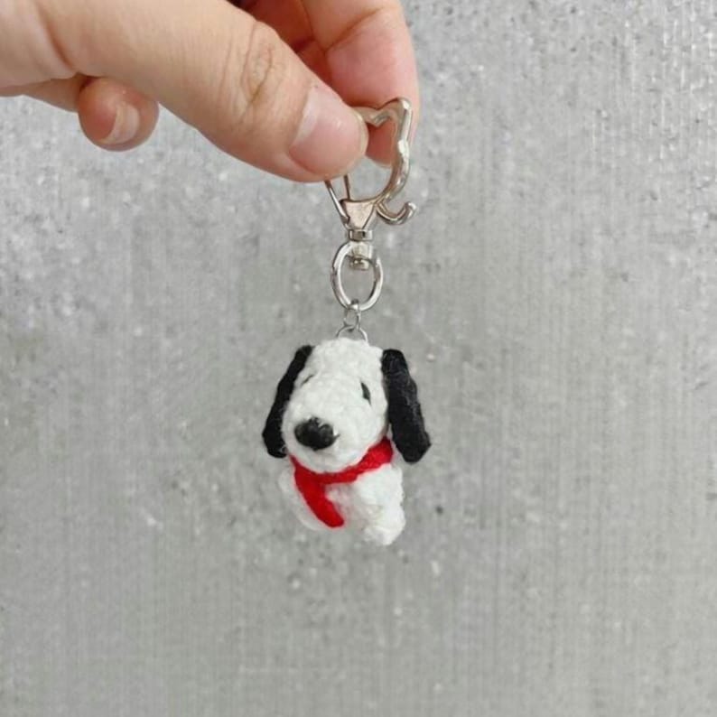 K&ouml;nnte beinhalten: Ein wei&szlig;-schwarzer Snoopy-Schl&uuml;sselanh&auml;nger mit rotem Schal. Der Schl&uuml;sselanh&auml;nger besteht aus einem weichen, strukturierten Material und hat einen silberfarbenen Verschluss. Die Snoopy-Figur hat schwarze Ohren und eine schwarze Nase.