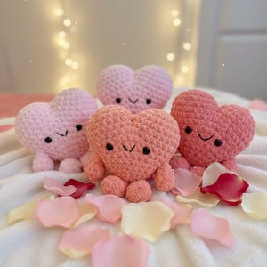 May include: Four handmade crochet heart-shaped plush toys in shades of pink and coral, each with embroidered eyes and a smiling mouth. The toys are arranged on a white surface with scattered rose petals.