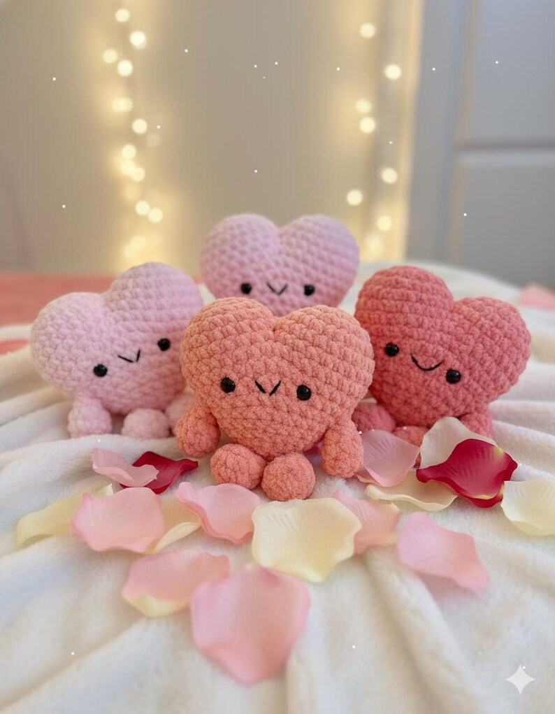 May include: Four handmade crochet heart-shaped plush toys in shades of pink and coral, each with embroidered eyes and a smiling mouth. The toys are arranged on a white surface with scattered rose petals.
