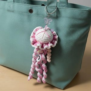 May include: A handmade crochet jellyfish keychain in white and pink, with a butterfly charm. The jellyfish has a round head and long, curly tentacles. It is hanging from a silver keyring attached to a teal tote bag.