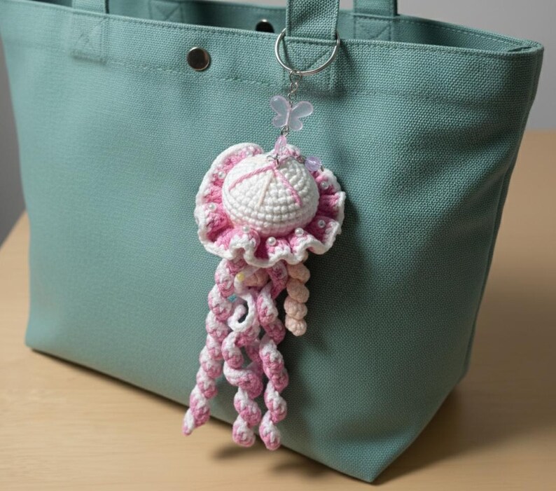 May include: A handmade crochet jellyfish keychain in white and pink, with a butterfly charm. The jellyfish has a round head and long, curly tentacles. It is hanging from a silver keyring attached to a teal tote bag.