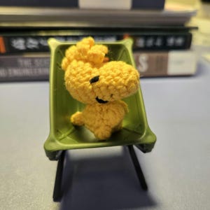 K&ouml;nnte beinhalten: Eine handgefertigte gelbe H&auml;kelfigur von Woodstock aus dem Peanuts-Comic, die in einem kleinen gr&uuml;nen Plastikstuhl sitzt. Die Figur hat einen schwarzen, l&auml;chelnden Mund und Augen. Der Stuhl hat schwarze Beine und einen gr&uuml;nen Sitz.