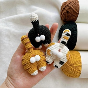 Op de afbeelding: Drie handgemaakte gehaakte kattenkontornamenten in zwart, oranje en wit met pluizige witte pompons. De kattenkonten worden in een hand gehouden, met garen op de achtergrond. De ornamenten zijn een leuk, grillig ambacht.