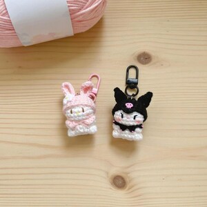 May include: Two handmade crochet keychains. One is a pink bunny with a flower, the other is a black cat with a skull. Both are white and black. The keychains are on a wooden surface with a pink yarn ball in the background.