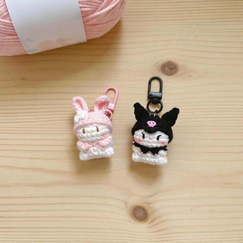 May include: Two handmade crochet keychains. One is a pink bunny with a flower, the other is a black cat with a skull. Both are white and black. The keychains are on a wooden surface with a pink yarn ball in the background.