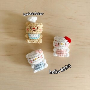 May include: Three handmade crochet figures: a bear wearing a chef's hat and apron, and two Hello Kitty figures. The bear is tan with a white hat and blue apron. The Hello Kitty figures are white with pink and blue accents.