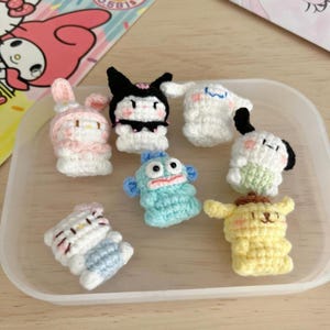 May include: A collection of seven small, crocheted character figures in a clear plastic container. The figures include a pink bunny, a black and white character, a white ghost, a blue fish, a white dog, a yellow dog, and a white cat.