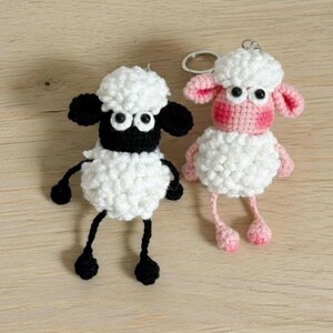 May include: Two handmade crochet sheep keychains. One sheep has black ears, face, and legs, and the other has pink ears, face, and legs. Both have white fluffy bodies and large googly eyes. The keychains are on a light brown wooden surface.
