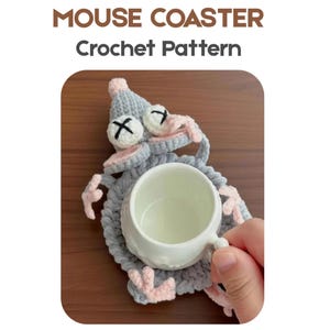 Puede incluir: Un posavasos de ratón de ganchillo gris y rosa con una taza de cerámica blanca. El posavasos tiene un diseño de cara de ratón con ojos negros en forma de cruz y detalles en rosa. El texto "MOUSE COASTER Crochet Pattern" está en la parte superior.