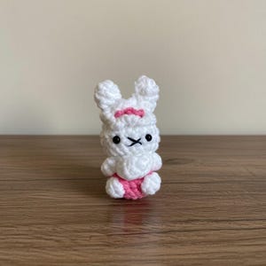 May include: A small, handmade crochet bunny in white with pink accents. The bunny has black eyes and a stitched mouth. A pink bow is on its head, and pink shorts are on its lower body. The bunny is sitting on a wooden surface.
