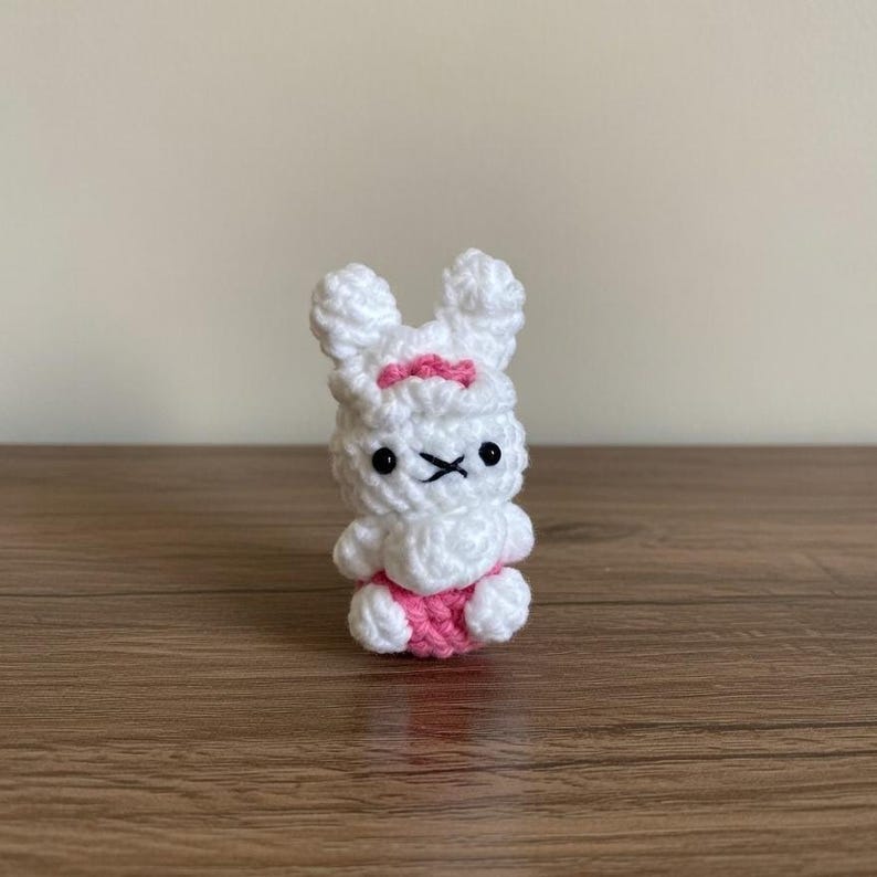 May include: A small, handmade crochet bunny in white with pink accents. The bunny has black eyes and a stitched mouth. A pink bow is on its head, and pink shorts are on its lower body. The bunny is sitting on a wooden surface.
