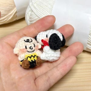 K&ouml;nnte beinhalten: Handgefertigte H&auml;kelfiguren von Charlie Brown und Snoopy. Charlie Brown tr&auml;gt ein gelbes Hemd mit einem schwarzen Zickzackmuster. Snoopy ist wei&szlig; mit schwarzen Ohren und einem roten Kragen.