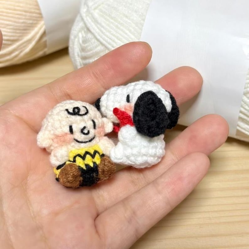 K&ouml;nnte beinhalten: Handgefertigte H&auml;kelfiguren von Charlie Brown und Snoopy. Charlie Brown tr&auml;gt ein gelbes Hemd mit einem schwarzen Zickzackmuster. Snoopy ist wei&szlig; mit schwarzen Ohren und einem roten Kragen.