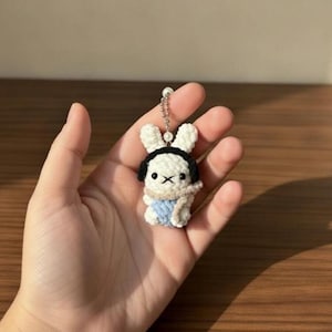 May include: A handmade crochet bunny keychain. The white bunny has black accents, a black headband, and a blue scarf. The keychain is attached to a silver chain with a pearl bead.