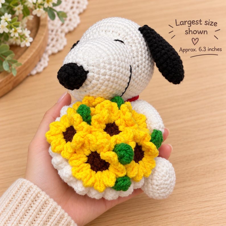 May include: A white and black crocheted dog toy holding a bouquet of yellow sunflowers with brown centers and green leaves. The toy features a red collar and text stating Largest size shown Approx. 6.3 inches.