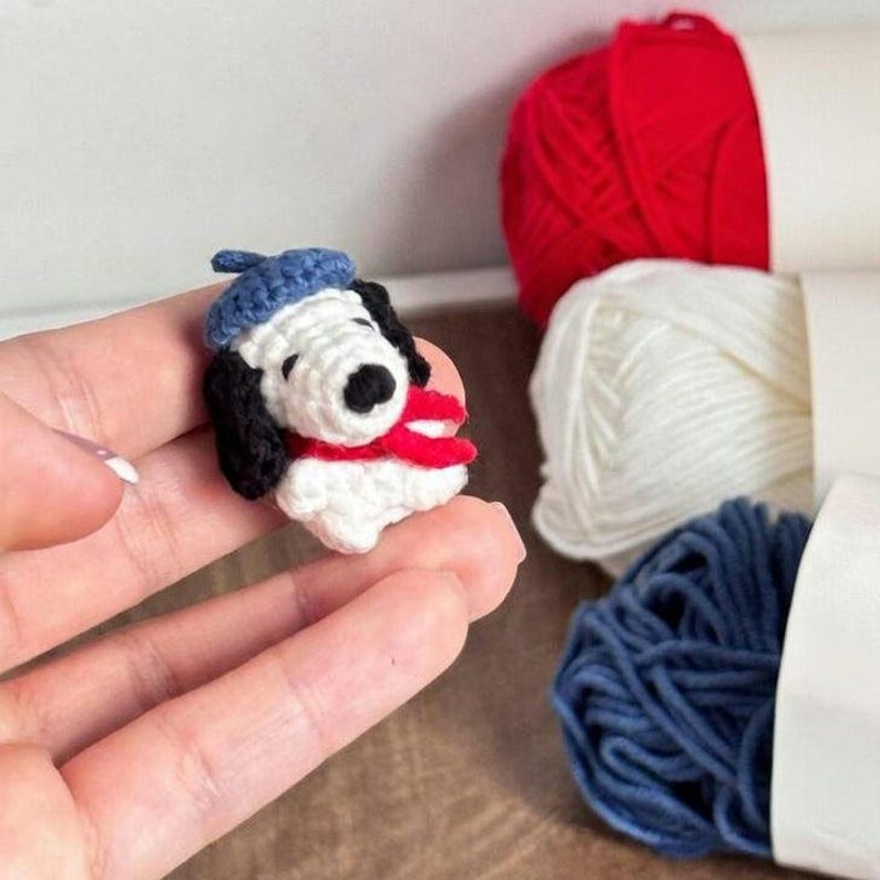 K&ouml;nnte beinhalten: Eine handgefertigte Snoopy-Figur aus H&auml;kelarbeit, die eine blaue Baskenm&uuml;tze und einen roten Schal tr&auml;gt. Die Figur ist wei&szlig; mit schwarzen Ohren und einer schwarzen Nase. Der Hintergrund umfasst rote, wei&szlig;e und blaue Garnkn&auml;uel.