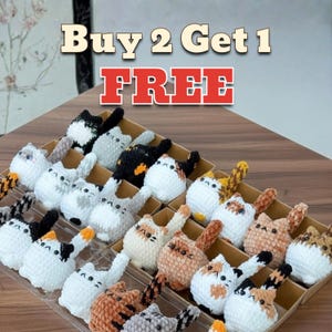 May include: A display of crocheted cat figurines in various colors, including white, gray, black, and orange. Each cat has a unique pattern and is arranged in a tiered display. The text "Buy 2 Get 1 FREE" is displayed at the top.