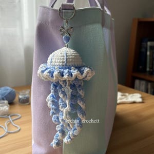 May include: A handmade crochet jellyfish charm in white and light blue, hanging from a silver-tone clasp on a pastel striped tote bag. The jellyfish has a ruffled edge and dangling tentacles. The text "@chin_crochett" is visible.