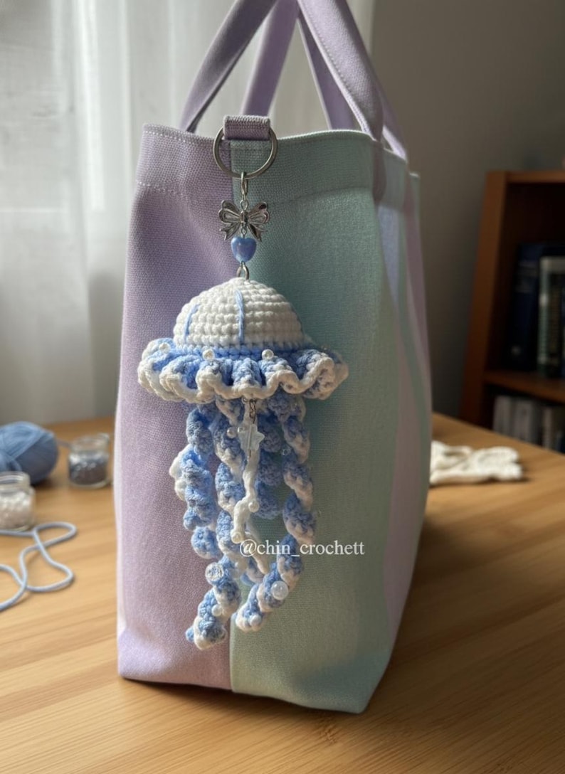 May include: A handmade crochet jellyfish charm in white and light blue, hanging from a silver-tone clasp on a pastel striped tote bag. The jellyfish has a ruffled edge and dangling tentacles. The text "@chin_crochett" is visible.