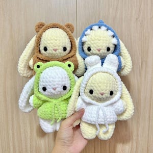 May include: Four handmade crochet bunny dolls in various outfits. One bunny wears a brown bear hoodie, another a blue shark hoodie, a third a green frog hoodie, and the last a white hoodie. Each bunny has black eyes and long ears.