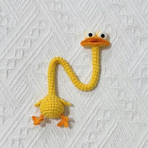 May include: A handmade yellow crochet duck with a long, curved neck, orange beak, and feet. The duck has large, googly eyes and is lying on a white, textured surface. This whimsical item is perfect for a nursery or as a decorative piece.