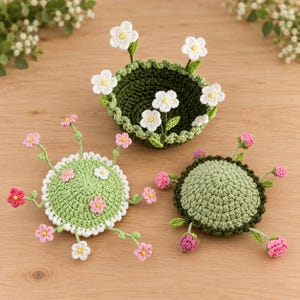 May include: Three round, crocheted items featuring floral designs. One is a dark green bowl with white flowers. Two are light green, circular, flat cushions adorned with small pink and white crocheted flowers on thin, protruding stems.