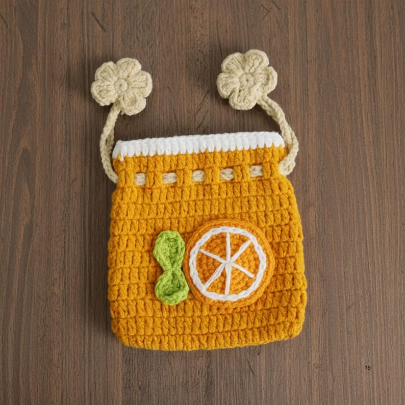 K&ouml;nnte beinhalten: Eine handgefertigte H&auml;keltasche in leuchtendem Gelb, mit wei&szlig;em Besatz und zwei beigen Blumenakzenten. Die Tasche ist mit einer gr&uuml;nen Schleife und einer geh&auml;kelten Orangenscheibe verziert. Die Tasche hat einen beigen Riemen.