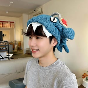 以下が含まれることがあります： 白い歯、単眼、赤と白のアクセントが付いた、漫画のサメの形をした青いかぎ針編みの帽子。帽子の上部には灰色のループがあり、人がかぶっています。