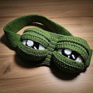 Puede incluir: Una máscara para dormir de ganchillo verde hecha a mano con la forma de una cara de rana de dibujos animados con grandes ojos expresivos. Los ojos son blancos con pupilas negras, y la máscara tiene una banda ancha y cómoda para un uso seguro.