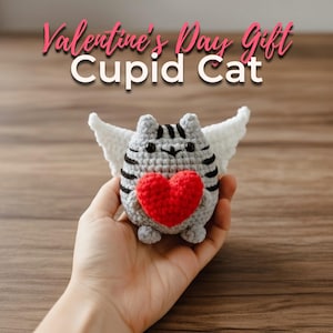 May include: A crocheted gray cat with black stripes, white wings, and a red heart. The text "Valentine's Day Gift Cupid Cat" is displayed above the cat. The cat is held in a hand.