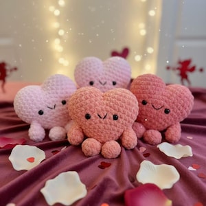 May include: Four crocheted heart-shaped plush toys in varying shades of pink and coral, each with a smiling face and small legs. The hearts are arranged on a deep red velvet surface scattered with rose petals and heart-shaped confetti.