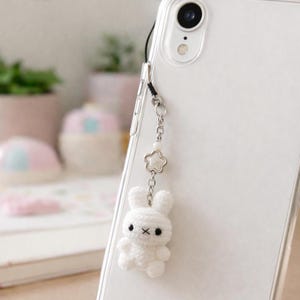 May include: A white crochet bunny phone charm with black eyes and a small X for a mouth. The charm hangs from a silver chain with a star-shaped charm and a pearl bead. The charm is attached to a clear phone case.