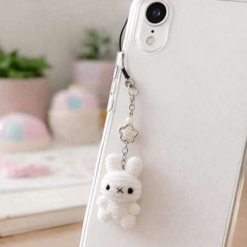 May include: A white crochet bunny phone charm with black eyes and a small X for a mouth. The charm hangs from a silver chain with a star-shaped charm and a pearl bead. The charm is attached to a clear phone case.