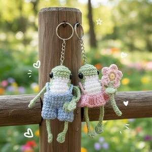 May include: Two handmade crochet frog keychains. One frog wears blue overalls, the other a pink dress and holds a flower. Both have green heads, white bellies, and black eyes. They hang from silver keyrings.