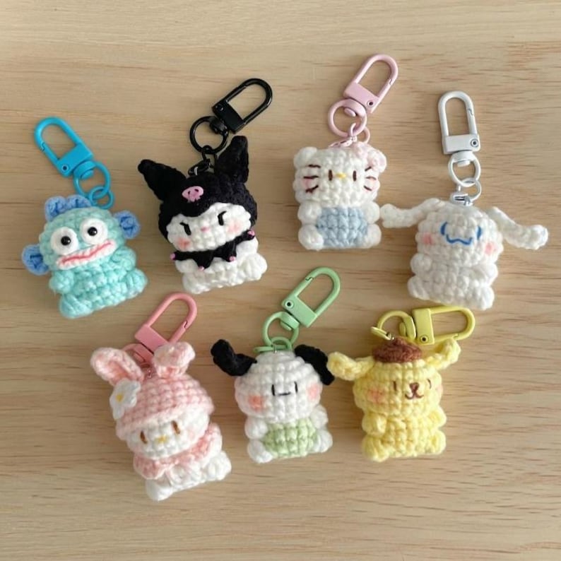 May include: A collection of handmade crochet keychains featuring various cartoon characters. The keychains are in various colors, including blue, black, white, pink, and yellow, and each has a metal clasp for attachment.