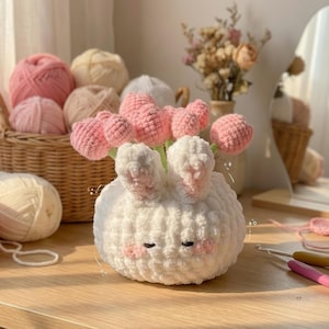 May include: A handmade white crochet bunny vase with pink accents, holding pink crochet tulips. The bunny has long ears and closed eyes. A basket of yarn balls in various shades of pink and cream is in the background.