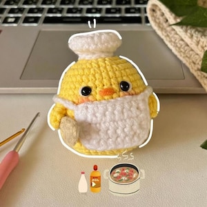 Può includere: Un pulcino giallo all'uncinetto fatto a mano con cappello da chef, grembiule e mascherina bianchi. Il pulcino tiene in mano un oggetto beige, forse un cucchiaio o una spatola. L'immagine include un laptop, uncinetti e illustrazioni a tema cibo.