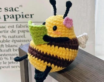 May include: Crocheted bee figurine with yellow and brown stripes, black antennae, and a pink flower. The bee holds a green book and has white wings. The bee is sitting on a wooden surface.