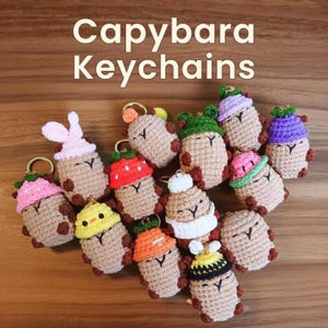 May include: A collection of crocheted capybara keychains, each with a unique hat design. The keychains are in various colors, including pink, green, yellow, and orange. The text "Capybara Keychains" is displayed at the top of the image.