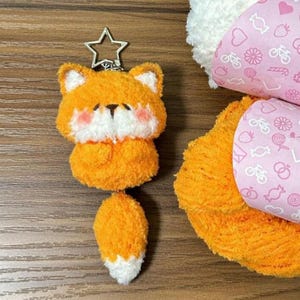 May include: A fluffy orange fox keyring with white detailing on the ears and chest, and a white-tipped tail. The fox has pink cheeks and a black nose and eyes. A silver star-shaped charm is attached to the top.