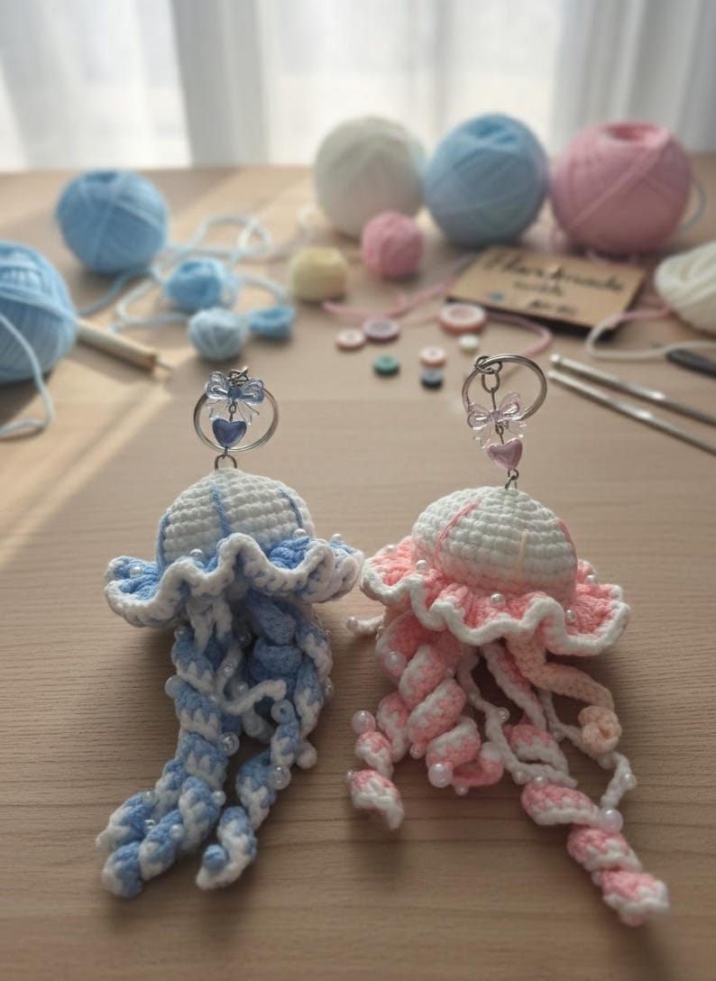 May include: Two handmade crochet jellyfish keychains, one blue and one pink, with white accents and pearl-like beads. Each keychain has a silver ring and a decorative charm. The background includes yarn balls and crafting supplies.