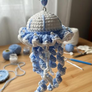 May include: A handmade crochet jellyfish keychain in white and light blue. The jellyfish has a ruffled edge and dangling tentacles, adorned with small white beads. A silver-tone bow and heart charm are attached to the top.