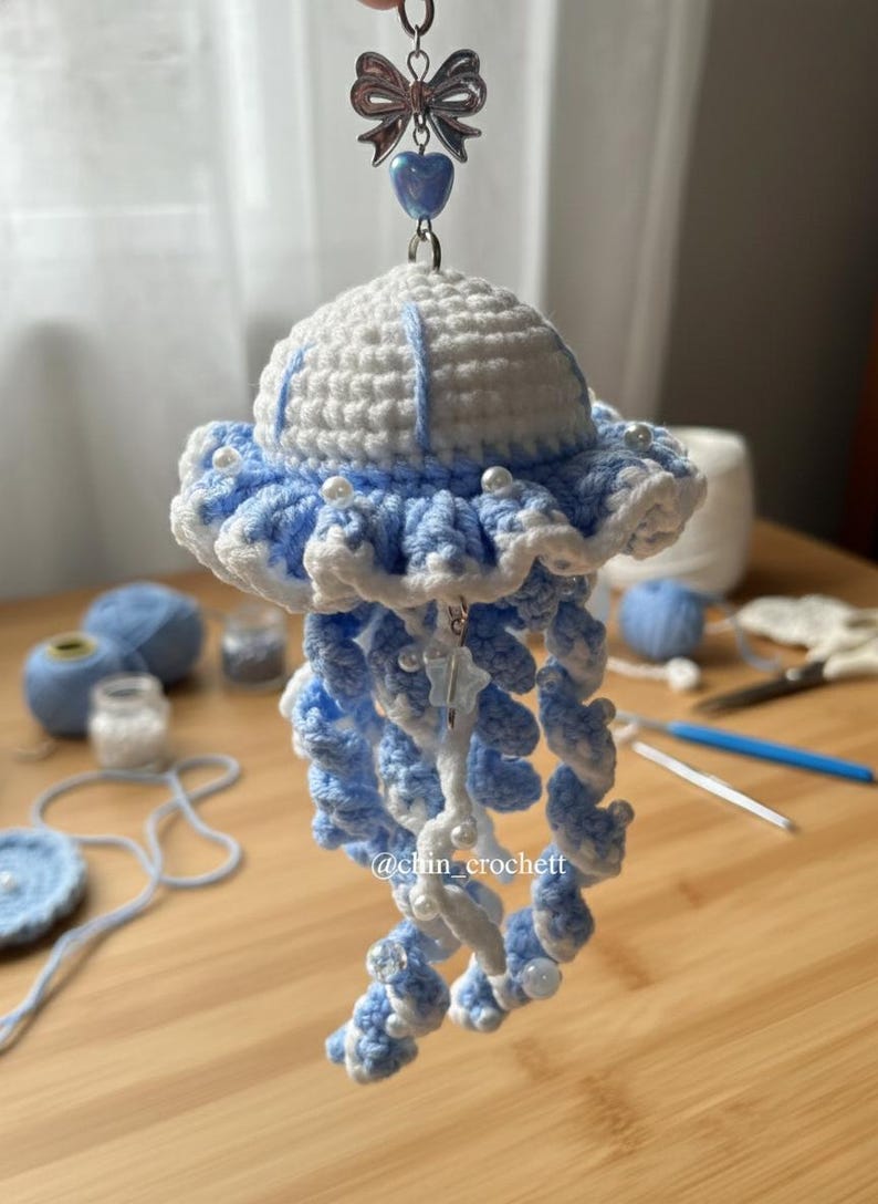 May include: A handmade crochet jellyfish keychain in white and light blue. The jellyfish has a ruffled edge and dangling tentacles, adorned with small white beads. A silver-tone bow and heart charm are attached to the top.
